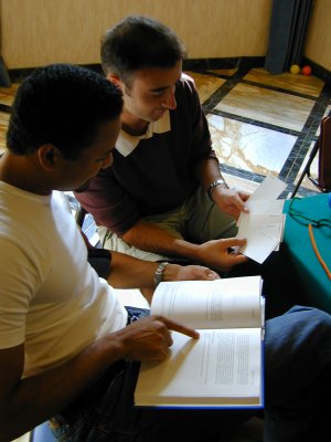 One person looking at cards, sitting next to a person pointing at a page in a book