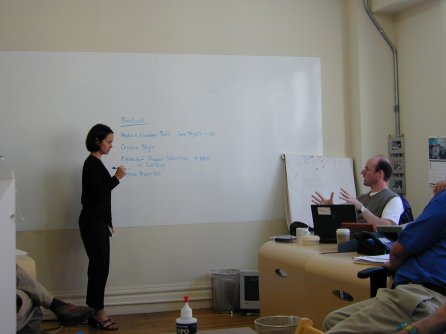 team sits in a circle planning, woman at white board writes notes down